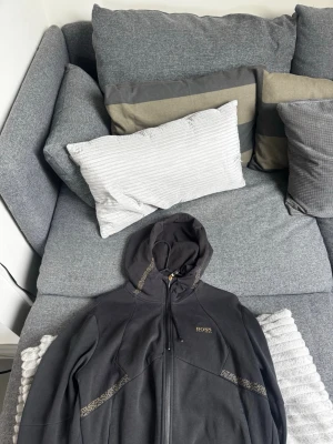Svart hoodie från BOSS med mönster - Säljer en svart hoodie från BOSS med guldfärgade detaljer och mönster längs dragkedjan och fickorna. Hoodien har huva med snörning och är tillverkad i mjukt material som känns skönt mot huden. Perfekt för dig som vill ha en stilren och exklusiv look. Pris diskuteras