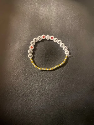 Pärlarmband med text och hjärtan - Fint armband med vita runda pärlor som stavar ut 'EN DAG I TAGET' och små röda hjärtan. Resten av armbandet består av små gula och vita pärlor. Perfekt accessoar för dig som gillar personliga smycken.