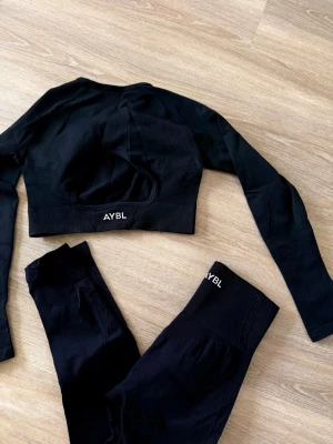 Svart seamless set från AYBL - Säljer ett svart seamless set från AYBL med långärmad crop top och matchande tights. Setet har diskreta ribbade detaljer och är tillverkat i stretchigt material som sitter skönt mot kroppen. Perfekt för dig som gillar stilrena träningskläder med snygg passform.