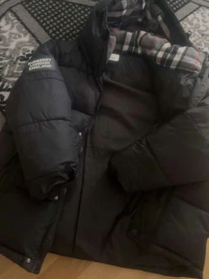 Svart pufferjacka från Burberry - Svart pufferjacka från Burberry med avtagbara ärmar som gör den till en väst. Jackan har en huva med klassiskt rutigt foder och två dragkedjefickor framtill. Patch med Burberry London England på ärmen. Perfekt för kalla vinterdagar.