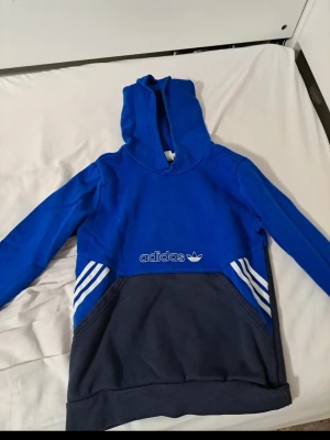 Adidas hoodie - Snygg hoodie från Adidas i blå och mörkblå färg med klassiska vita ränder på sidorna och Adidas-logga framtill. Hoodien har stor huva och magficka. Tillverkad i mjukt bomullsmaterial, perfekt för en avslappnad streetwear-look.jätte snygg size 158 cm priset är 150 kr 