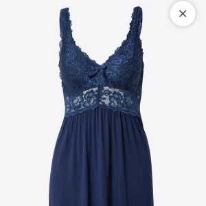 Mörkblå spetsnattlinne med rosett - Snyggt nattlinne i mörkblått med breda axelband och vacker spets upptill. Dekorativ rosett framtill och genombruten spets över bysten och midjan. Kjoldelen är mjukt fallande i ett stretchigt tyg. Perfekt för en mysig kväll hemma.