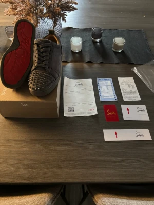 Christian Louboutin gråa sneakers med nitar - Säljer ett par svarta sneakers från Christian Louboutin med klassiska röda sulor och coola nitar över tån. Skorna är låga och har snörning samt är tillverkade i mocka för en lyxig känsla. Perfekta för dig som vill sticka ut med din stil. Skorna har en liten defekt på bitarna som syns på bild nr.4 men det är inget som man lägger märke till vid användning. Tveka inte på att höra av dig// Mvh Liam.