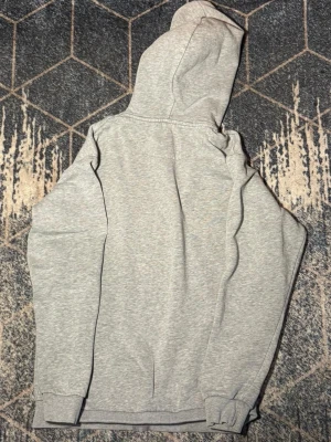 Ljusgrå hoodie från Jack & Jones - Ljusgrå hoodie från Jack & Jones med dragkedja och huva. Tillverkad i mjukt och bekvämt material, perfekt för chill dagar. Klassisk passform med ribbade muddar vid ärmslut och nederkant. Enkel och stilren design utan tryck.
