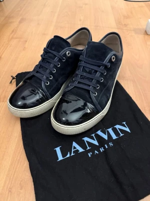 Mörkblå sneakers från Lanvin - Snygga mörkblå sneakers från Lanvin med ovandel i mocka och lackad tå. Skorna har vita sulor, mörkblå skosnören och metallfärgade öljetter. Klassisk låg modell som ger en clean och exklusiv vibe. Perfekta för dig som gillar stilrena detaljer. Dustbag ingår. Snabb affär ger billigare pris, vill få sålt snabbt.