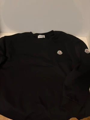 Svart sweatshirt från Moncler - Snygg svart sweatshirt från Moncler med rund halsringning och klassisk logotyp på bröstet. Tröjan har en enkel och stilren design, perfekt för dig som gillar exklusiva streetwear-plagg. Tillverkad i mjukt material för skön komfort.