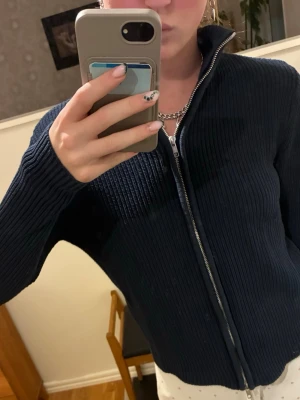 Mörkblå ribbad ziptröja - Säljer en mörkblå ribbstickad tröja med dragkedja framtill och hög krage. Tröjan har en clean look och är perfekt för lager på lager. Materialet ser ut att vara mjukt och stretchigt, vilket gör den bekväm att bära.