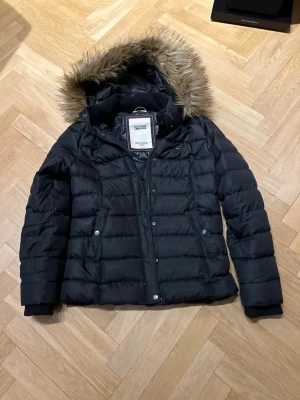 Svart dunjacka Hilfiger Denim M - Svart dunjacka från Hilfiger Denim med avtagbar huva och fuskpäls. Jackan har quiltad design, dragkedja och tryckknappar framtill samt två fickor med lock. Perfekt för kalla vinterdagar och riktigt snygg streetstil.
