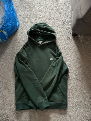Mörkgrön hoodie från Lacoste - Snygg mörkgrön hoodie från Lacoste med klassisk krokodillogga på bröstet. Hoodien har huva och känguruficka framtill. Tillverkad i mjukt material som känns skönt mot huden. Perfekt för en avslappnad och stilren look.