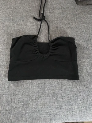 Svart bandeau topp med knyt - Snygg svart bandeau topp med knyt i nacken och rynkad detalj framtill. Perfekt till sommarens outfits och enkel att matcha med olika stilar. Mjuk och stretchig känsla.