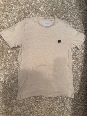 Beige t-shirt från Les Deux - Snygg beige t-shirt från Les Deux med klassisk passform och korta ärmar. T-shirten har en liten svart patch med romerska siffran II på bröstet. Tillverkad i mjuk bomull som känns skön mot huden. Perfekt till jeans eller shorts.