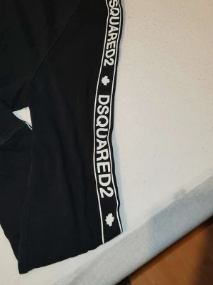 Svart loungewear-set från Dsquared2 - Snyggt svart loungewear-set från Dsquared2 med vita logotexter och kanter längs ärmar och ben. Setet har en avslappnad passform och är gjort i mjukt bomullsmaterial, perfekt för chill dagar hemma. Klassisk crewneck och elastiska muddar för extra komfort.