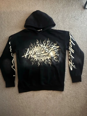 Svart hoodie Hellstar Records flames - Svart hoodie från Hellstar Records med stort flames-tryck i beige och vitt på bröstet och ärmarna. Hoodien har huva och magficka, samt coolt grafiskt tryck som ger streetwear-vibe. Perfekt för dig som gillar unika statement-plagg.