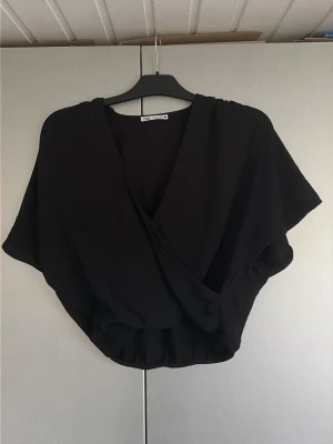 Svart omlottopp från Zara - Snygg svart omlottopp från Zara med korta ärmar och croppad passform. Toppen har en djup v-ringning och elastisk nederkant som ger en avslappnad men trendig look. Perfekt att matcha med höga jeans eller kjol.