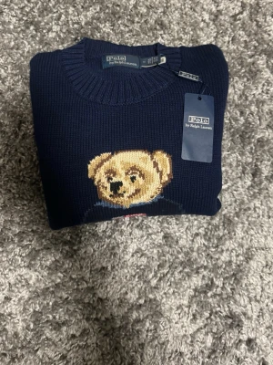 Mörkblå Polo Bear stickad tröja - Mörkblå stickad tröja från Polo Ralph Lauren med ikoniskt Polo Bear-motiv framtill. Tröjan har rund halsringning, ribbstickade muddar och RL-broderi nertill. Perfekt för dig som gillar klassisk och lekfull stil.