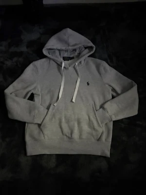 Grå hoodie från Polo Ralph Lauren - Snygg grå hoodie från Polo Ralph Lauren med klassisk huva och vita dragsnören. Tröjan har en känguruficka framtill och den ikoniska lilla loggan broderad på bröstet. Tillverkad i mjukt bomullsmaterial för maximal komfort.