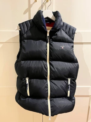 Svart dunväst från Gant - Snygg marinblå dunväst från Gant med vita dragkedjor och diskret logga på bröstet. Västen har hög krage, två fickor med dragkedja och quiltad design. Perfekt för lager på lager och ger en sportig vibe. Den har även en luva man kan gömma i nacken 