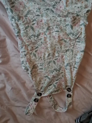 Blommig klänning från Mywear Young - Supersöt klänning med blommigt mönster i pastellfärger från Mywear Young. Hängselmodell med stora knappar framtill och en praktisk ficka på magen. Mjuk bomullskänsla och perfekt för en avslappnad stil.