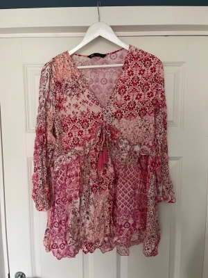 Blommig klänning från Zara - Romantisk kortklänning i rosa och rött med blommigt mönster och v-ringning. Klänningen har långa ärmar, snörning med dekorativa tofsar och volangkant. Tillverkad i ett lätt, luftigt material som passar perfekt för sommarens alla dagar.💗🌹