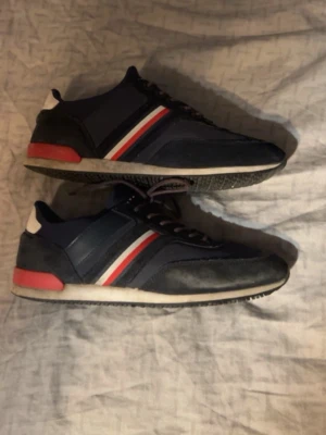  Tommy hilfiger Marinblå sneakers  - Snygga marinblå sneakers med röda och vita ränder på sidan samt röd detalj vid hälen. Skorna har lågt skaft och en mix av mocka och textilmaterial. Perfekta för dig som gillar sportig och stilren look.