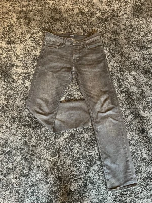 W29/L32 - Mörk grå tapered fit jeans från Jack & Jones - Säljer ett par mörk gråa tapered fit jeans från Jack & Jones med klassisk femficksdesign och snygga detaljer. Jeansen har raka ben och är tillverkade i mjukt denim-material med stretch för extra komfort. Perfekt för dig som gillar en stilren känsla.