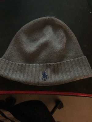 Grå mössa från Polo Ralph Lauren - Snygg grå mössa från Polo Ralph Lauren i mjuk och tvättbar merinoull. Klassisk ribbad kant och broderad blå logga framtill. Perfekt accessoar för att hålla värmen och addera stilpoäng till din outfit.