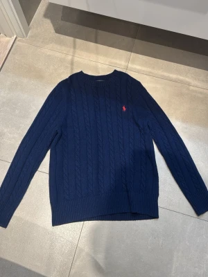 Mörkblå kabelstickad tröja Polo Ralph Lauren - Mörkblå kabelstickad tröja från Polo Ralph Lauren med rund halsringning och röd broderad logga på bröstet. Tröjan har långa ärmar och ribbstickade muddar vid ärmslut och nederkant. Perfekt för en klassisk och stilren look. 