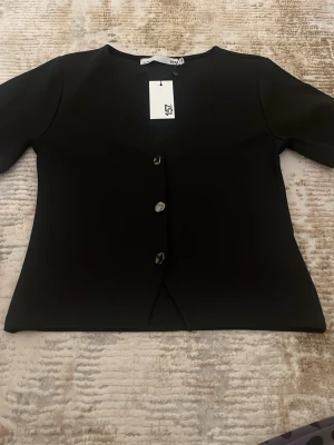 Svart kofta från 157 med knappar - Stilren svart kofta från 157 med tre dekorativa knappar framtill. Koftan har rund halsringning och halvlånga ärmar. Materialet är slätt och ger ett modernt intryck. Perfekt att matcha med jeans eller kjol för en enkel look.