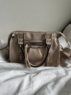 Beige handväska med gulddetaljer - Snygg beige handväska med guldfärgade metalldetaljer och svarta kontrastkanter. Väskan har både handtag och justerbar axelrem samt en liten ficka med dragkedja framtill. Perfekt storlek.