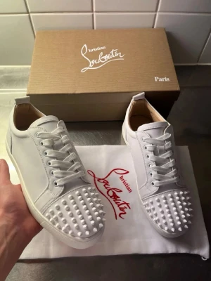 Vita Christian Louboutin sneakers med nitar - Snygga vita sneakers från Christian Louboutin i läder med ikoniska röda sulor och coola vita nitar på tån. Klassisk låg modell med vita skosnören och exklusiv känsla. Perfekta för dig som vill sticka ut med stil.