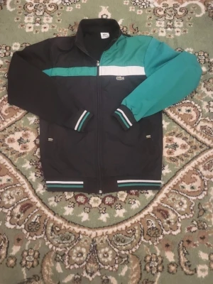 Svart och grön hoodie från Lacoste - Snygg hoodie från Lacoste i svart med gröna och vita detaljer. Dragkedja framtill, ribbade muddar med randigt mönster och två sidofickor. Klassisk Lacoste-logga på bröstet och cool färgblockad design. Perfekt för dig som gillar streetwear och sportig stil.