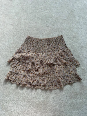 Volangkjol från Neo Noir - Supersöt kort kjol med blommigt mönster i beige, brunt och lila. Kjolen har smock i midjan och flera volanglager som ger en flowy känsla. Perfekt för dig som gillar romantisk och bohemisk stil. Denna kjol är en av mina favoriter och jag har använt den sparsamt. 💕Nypris ca 600 kr