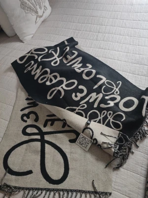 Svart och beige sjal från Loewe - Snygg sjal från Loewe i svart och beige med grafiskt mönster och fransar i kanterna. Sjalen har en lyxig känsla och är tillverkad i Frankrike. Perfekt accessoar för att lyfta din outfit med en trendig touch.