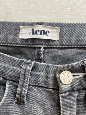Grå jeans från Acne Studios - Snygga grå jeans från Acne Studios med klassisk femficksmodell och silverfärgad knapp med logga. Jeansen har en cool tvätt och detaljer som läderpatch med Acne-logga bak. Perfekta för en trendig och avslappnad stil. Storlek 30/32 