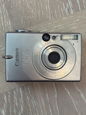 Canon Digital IXUS II kompaktkamera - Snygg och smidig digitalkamera från Canon, modell IXUS II. Kameran är silverfärgad med rektangulär form och har ett robust hölje i metall. Utrustad med inbyggd blixt, liten LCD-skärm och klassisk knappnavigering. Användarhandbok, laddare och sladd inkluderad!