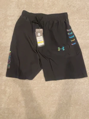Svarta träningsshorts från Under Armour - Svarta träningsshorts från Under Armour med färgglada detaljer och logga på ena benet. Shortsen har elastisk midja med snörning och är tillverkade i ett lätt och snabbtorkande material. Perfekta för sport eller gym.