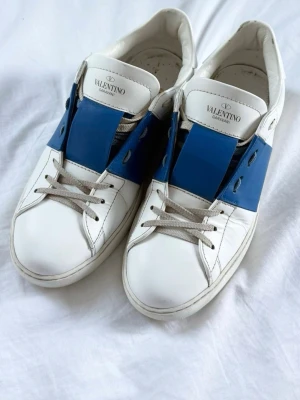 Valentino Garavani Rockstud - Snygga sneakers från Valentino Garavani i vitt läder med bred blå rem över vristen och vita skosnören. Klassisk låg modell med rund tå och diskret logga på plösen. Medföljer originalpåse och extra skosnören.