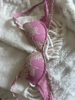 Rosa push-up bh med spetsdetaljer - Snygg rosa push-up bh med ljusbeige spetsdetaljer och vadderade kupor. Bh:n har justerbara axelband och fina spetskanter längs kuporna och banden. Materialet är mjukt och bekvämt, perfekt för dig som vill ha både stil och komfort.
