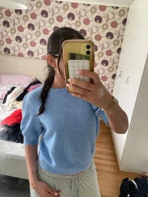 Ljusblå stickad topp med puffärm - Säljer en ljusblå stickad topp med korta puffärmar och ribbade kanter vid hals, ärmslut och nederkant. Tröjan har en mjuk och skön känsla, perfekt för lager på lager. Enkel och stilren design som passar till mycket.