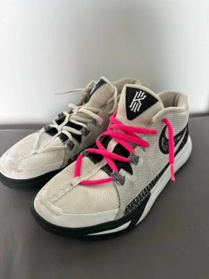 Kyrie basketskor - Snygga Nike Kyrie sneakers i vitt med svarta detaljer och olika färgade skosnören, ett par är rosa och det andra vitt. Skorna har mesh-ovandel och tydlig Kyrie-logga på plösen samt yttersula med bra grepp. Perfekta för dig som gillar basketstil och streetwear. Skick 6/10. Kom privat för mer info!