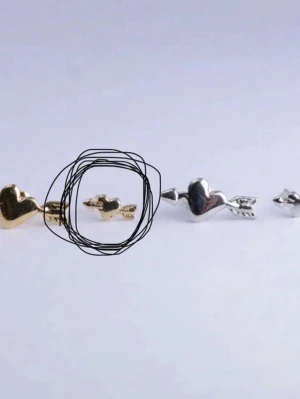 Örhänge hjärta med pil i guldton - Sött örhänge i form av ett hjärta med pil, i guldfärgad metall. Liten och stilren design som ger en lekfull touch till din look. Perfekt för dig som gillar unika detaljer och romantiska smycken.