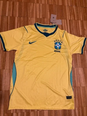 Brasilien fotbolls tröja,Gul  sporttröja  - Säljer en gul Brasilien vm tröja från  blå detaljer längs sidorna och på ärmsluten. Tröjan har korta ärmar och är tillverkad i ett lätt och ventilerande material som passar perfekt för träning och sportaktiviteter. Jag fick de som present och de va lite för stor för mig. De är riktigt bra kvalité.