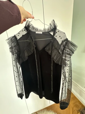 Svart meshblus med volanger och prickar - Trendig svart blus från NA-KD med transparenta meshärmar och volangdetaljer. Ärmarna har prickigt mönster och blusen har volanger vid axlar och hals. Perfekt för dig som gillar statement-plagg och snygga detaljer.