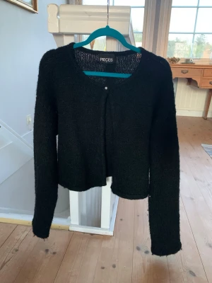 Svart kofta från Pieces - Säljer en svart, mjuk och lite fluffig kofta från Pieces. Koftan har rund halsringning, långa ärmar och en enkel knapp framtill. Perfekt att slänga över en topp för en chill look.