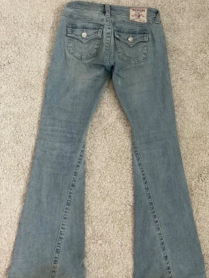 Ljusblå bootcut jeans True Religion ”joey” - Säljer ett par ljusblå bootcut jeans från True Religion med låg midja och klassiska fickor med lock och silverfärgade knappar bak. Jeansen har en snygg tvätt och är tillverkade i denim med lätt stretch för skön passform.