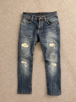 Nudie jeans  - Säljer ett par blå jeans från H&M med slitningar och hål på båda benen. Klassisk femficksmodell med normal passform och snygga detaljer. Perfekta för en avslappnad och trendig look.size 28:32 