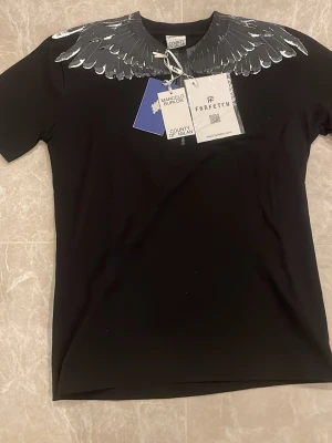Svart t-shirt med vingar från Marcelo Burlon - Snygg svart t-shirt från Marcelo Burlon med ikoniskt vingmönster i grått och vitt runt halsen. Klassisk passform och korta ärmar. Tillverkad i mjuk bomull som känns skön mot huden. Perfekt för dig som vill sticka ut med en unik detalj.