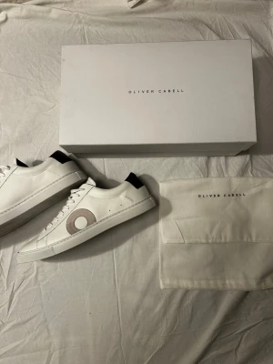 Vita sneakers från Oliver Cabell - Stilrena vita sneakers från Oliver Cabell med svart häl och en beige cirkeldetalj på sidan. Skorna har klassisk låg profil, vita snören och är tillverkade i slätt läder. Perfekta för en clean och modern look. Nypris ligger på 2500( när jag köpte dem) + 800 kr frakt från USA. Skriv vid minsta fundering.