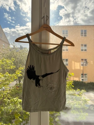 Grå linnetopp med örn och pärlor - Cool grå linnetopp med tryck av en svart örn och detaljerad pärlbroderi i form av en explosion med en liten dödskalle. Perfekt för dig som gillar unika detaljer och edgy stil.