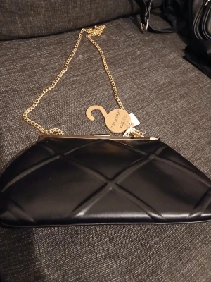 Svart kuvertväska med kedja Primark - Stilren svart kuvertväska från Primark med quiltat mönster och guldfärgad kedja. Väskan har en hård ram upptill och stängs med ett klassiskt metallspänne. Perfekt accessoar för att lyfta din outfit.